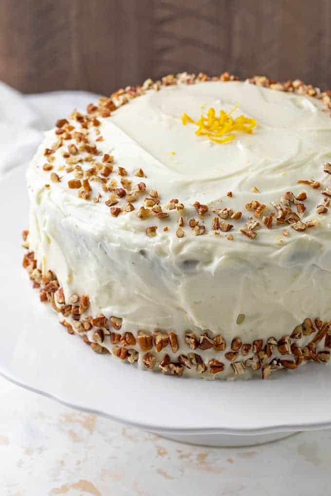 A round cake topped with chopped pecans and lemon zest on a round white cake stand.