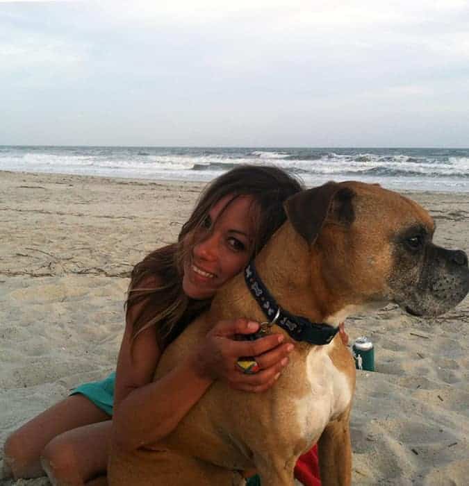 A young woman with brown hair hugging a boxer dog on the beach.