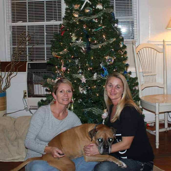 Two young women sitting in front of a Christmas tree with a boxer dog between them.
