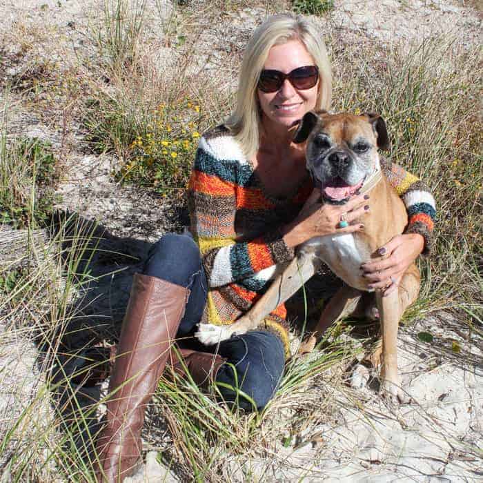 A woman with blonde hair and sunglasses sitting on the beach hugging a boxer dog.