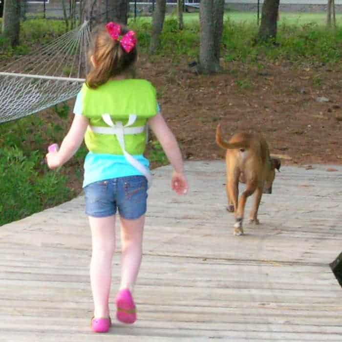 Back view of a little girl wearing a green life preserver and a boxer dog walking down a dock.