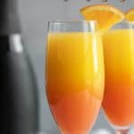 Two mimosas in champagne flutes. A black bottle of champagne is in the background. Overlay text at top of image.