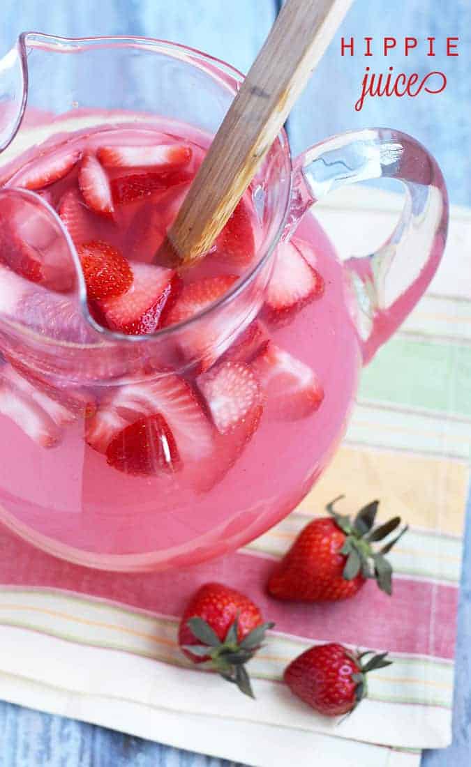 A glass pitcher filled with a pink beverage and sliced strawberries with a wooden spoon.
