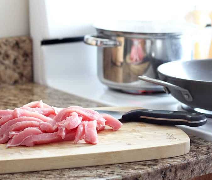 Raw pork sliced into strips on a cutting board on a counter by a stove.