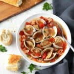 Steamed clams with smoky bacon and tomatoes in a white bowl and French bread on a wooden cutting board.