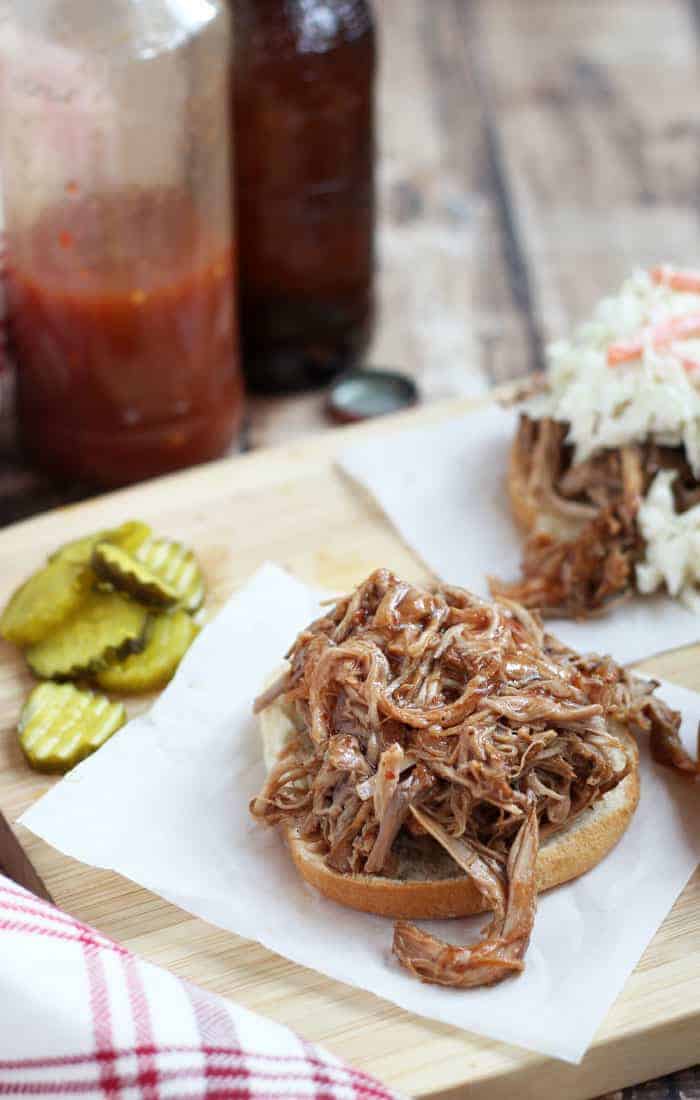 Pulled pork on an open faced hamburger bun beside sliced pickles on a wood surface.