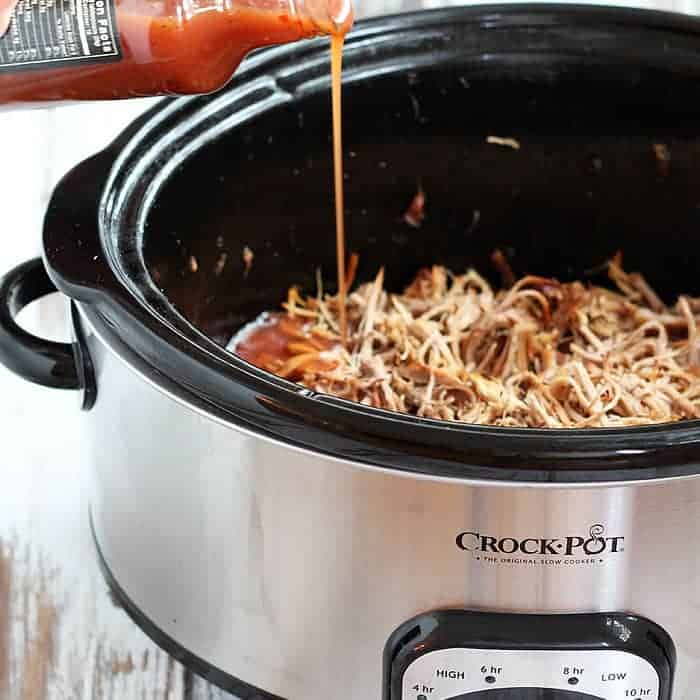 Pouring barbecue sauce over pulled pork in a stainless slow cooker.