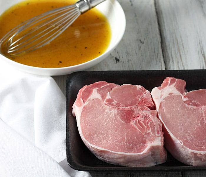 Two thick pork chops in a black foam tray. A bowl of marinade with a whisk is in the background.