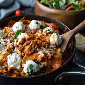 Lasagna in a large skillet with a wooden spoon and salad in a wooden bowl.