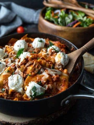 Lasagna in a large skillet with a wooden spoon and salad in a wooden bowl.