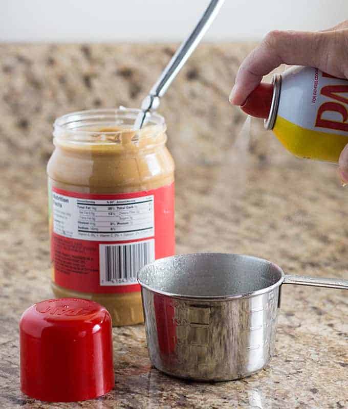 A measuring cup being sprayed with cooking spray beside a jar of peanut butter.