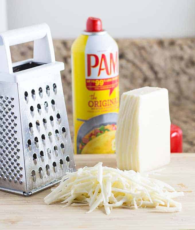 A cheese grater beside grated cheese and a can of cooking spray on a cutting board.