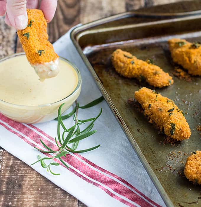 Dipping a breaded chicken finger in a bowl of dipping sauce beside a baking sheet with chicken fingers.