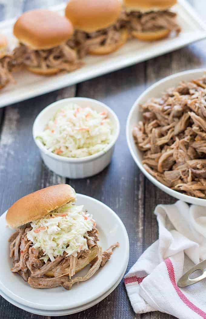 A shredded pork sandwich with coleslaw on a plate. A bowl of barbecue and coleslaw are in the background.