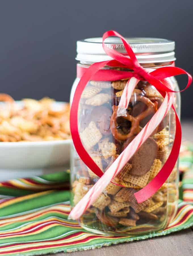 Chex snack mix in a jar with a red ribbon and candy cane tied around the rim. 