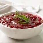 Front view of cranberry sauce garnished with a sprig of rosemary in a white bowl.