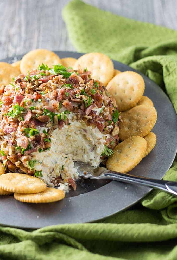 A cheese ball topped with bacon pieces surrounded by crackers on a silver plate.