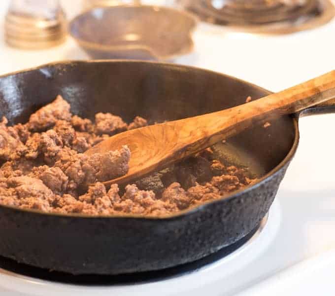 Browning ground beef in a skillet with a wooden spoon on a stove burner.