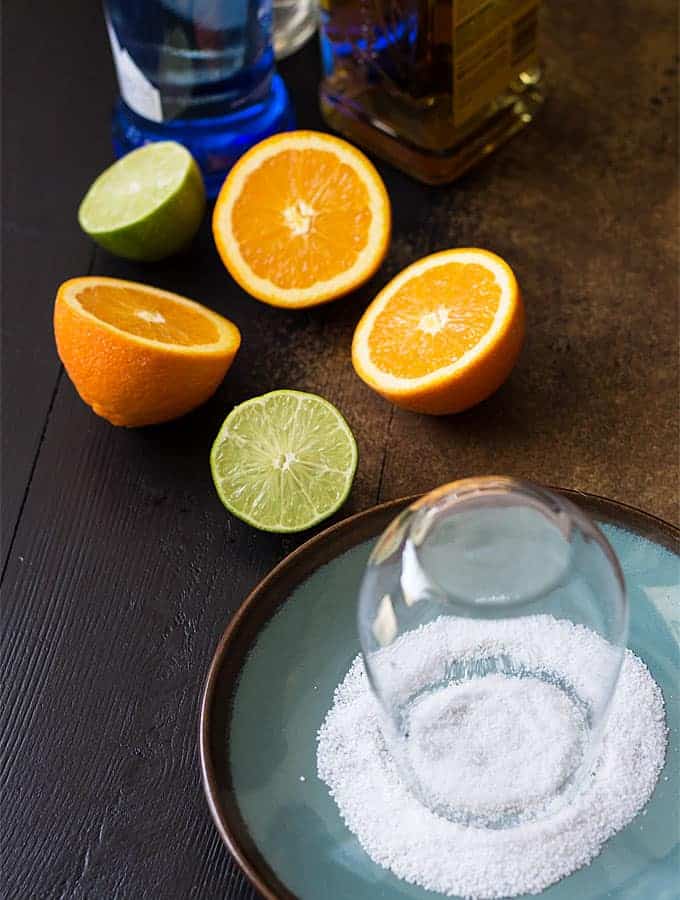 Overhead view of a glass being rimmed on a plate with salt, oranges and limes.