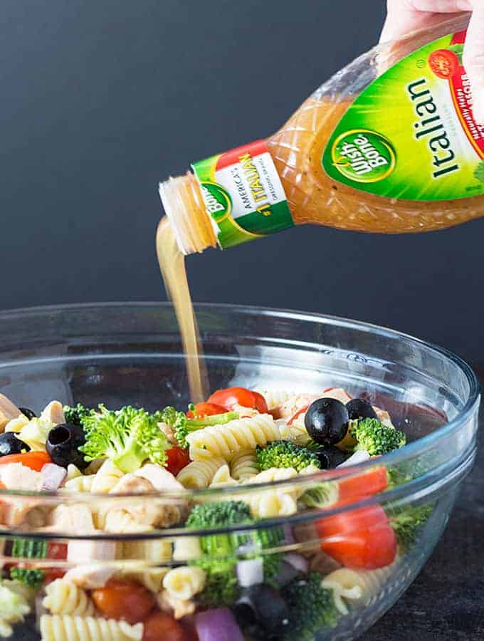 Pouring Italian dressing from a bottle into a bowl of pasta, chicken and vegetables.