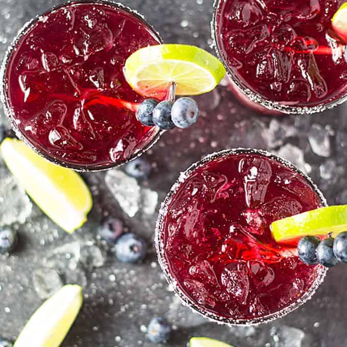 Overhead view of 3 blueberry margaritas on a dark surface with crushed ice on the surface.