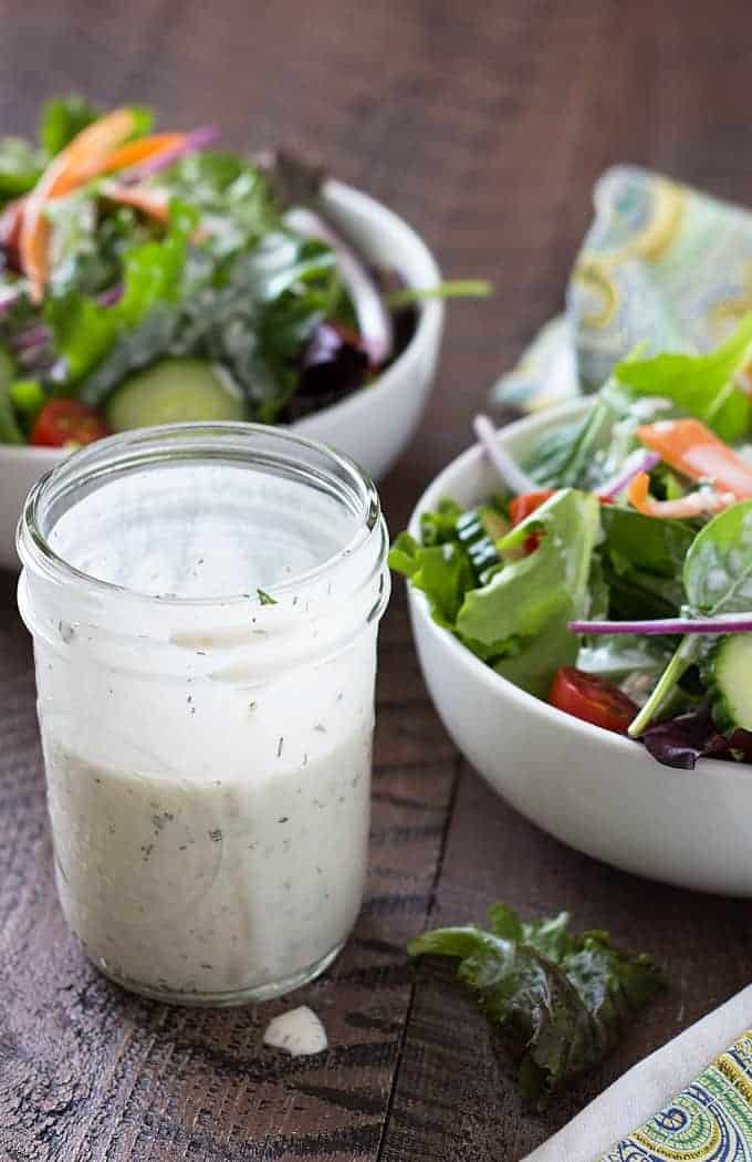 A jar of ranch dressing by two white bowls of salad on a wood surface.