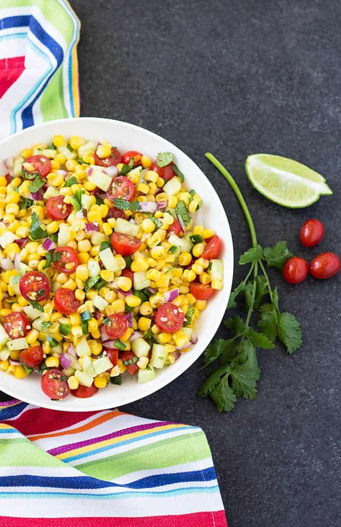 Overhead view of corn salad with cucumbers and tomatoes in a white bowl by a striped napkin.