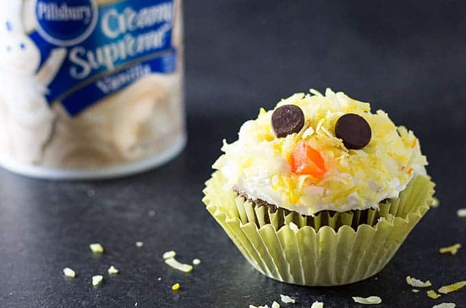 A cupcake decorated to resemble a baby chicken's face. A tub of frosting is in the background.