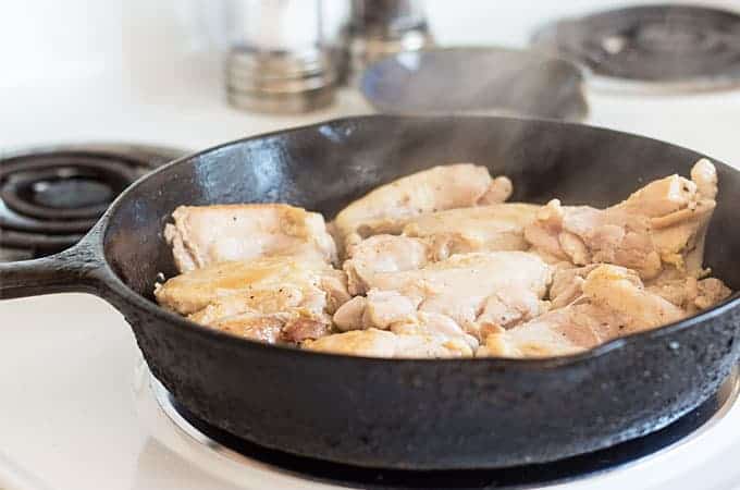 Chicken thighs being fried in a cast iron skillet on a stove burner.