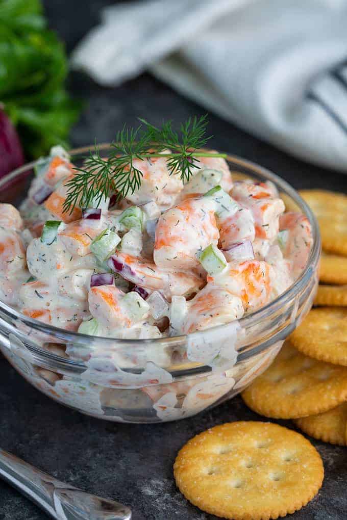 Shrimp salad garnished with fresh dill in a clear glass bowl beside crackers.
