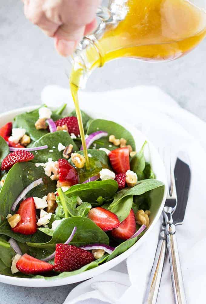 Pouring a vinaigrette over a strawberry and spinach salad in a white bowl.