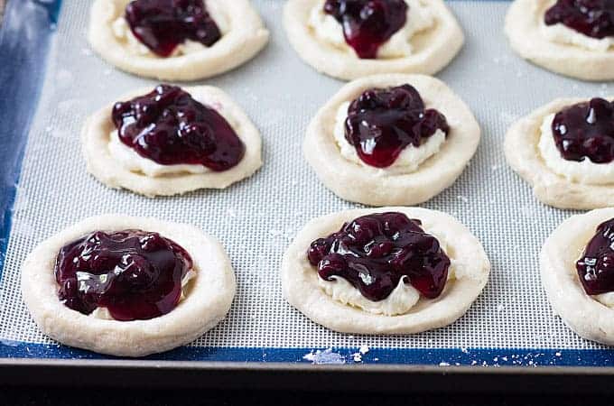 Canned biscuits that have been pressed in the center and filled with blueberry pie filling on a baking sheet.