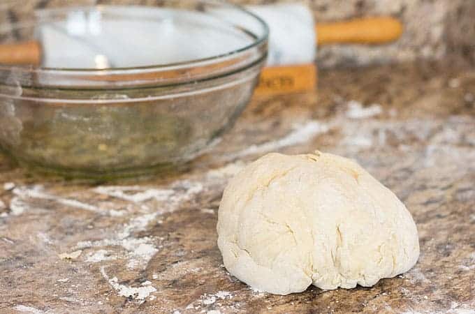 A ball of dough on a floured granite surface. A large bowl and rolling pin are in the background.