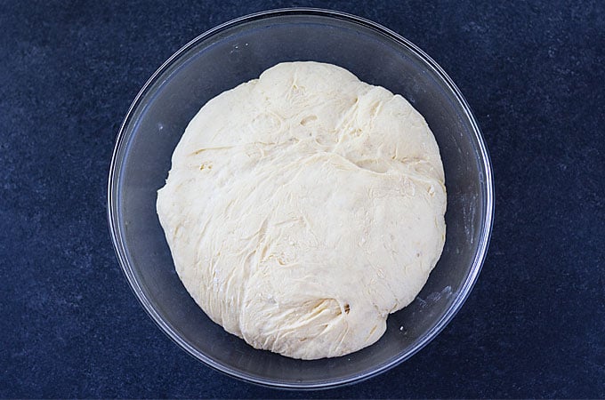 Overhead view of a ball of dough in a glass bowl on a dark surface.
