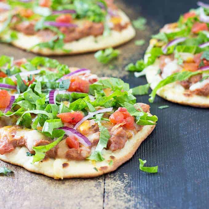 Closeup view of a flatbread pizza with refried beans, diced tomatoes, red onion, lettuce and cheese.