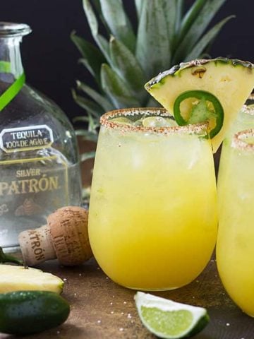 Three margaritas garnished with pineapple and jalapeno by a bottle of tequila.