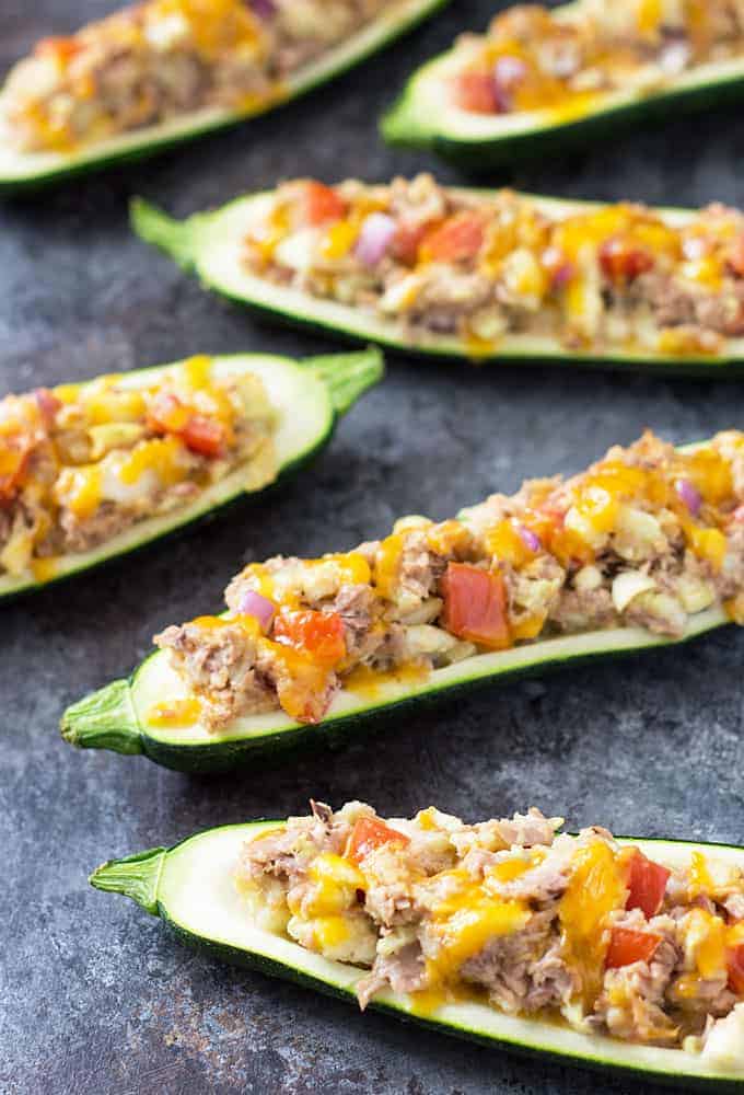 Six zucchini boats stuffed with a tuna, vegetable and cheese mixture on a dark surface. 