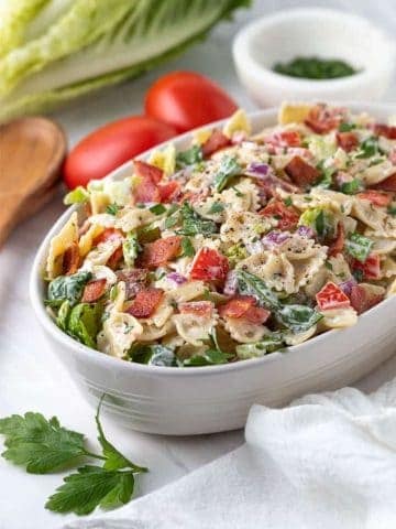 BLT Pasta Salad in an oval white serving bowl beside a wooden serving spoon and white kitchen towel.