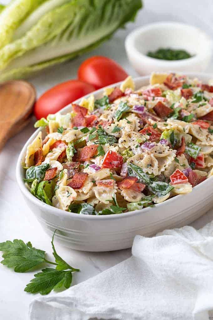 BLT Pasta Salad in an oval white serving bowl beside a serving spoon and white towel.