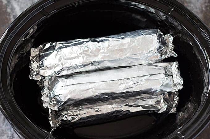 Three ears of corn wrapped in aluminum foil in a slow cooker insert.