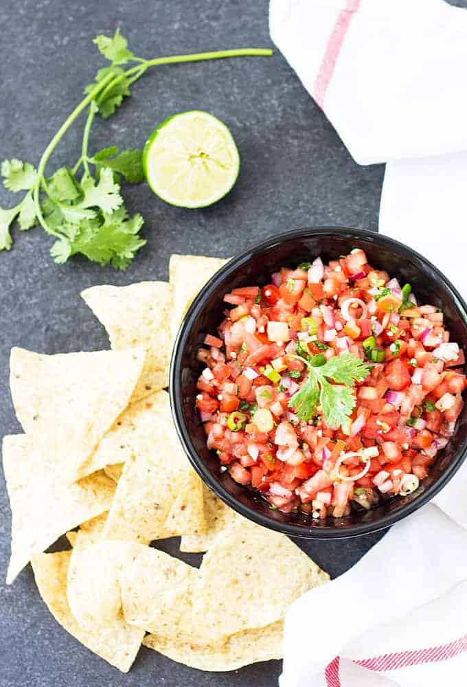 Overhead view of salsa in a black bowl beside tortilla chips, a sprig of cilantro and a lime.