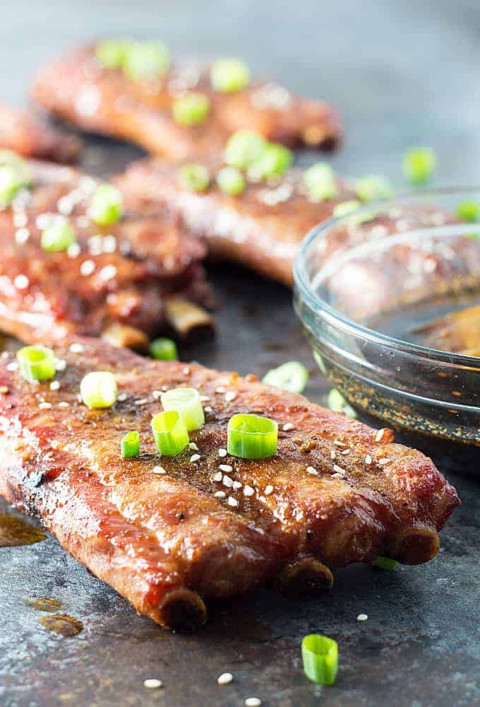 Grilled pork ribs topped with sliced green onions by a glass bowl of sauce.