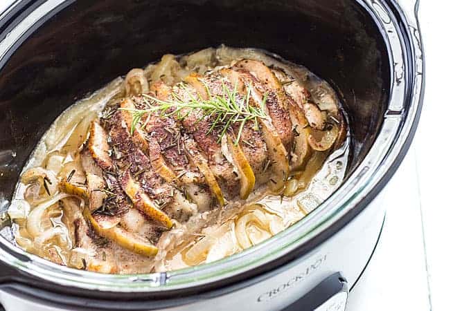 A cooked apple pork loin topped with a sprig of fresh rosemary in a slow cooker.