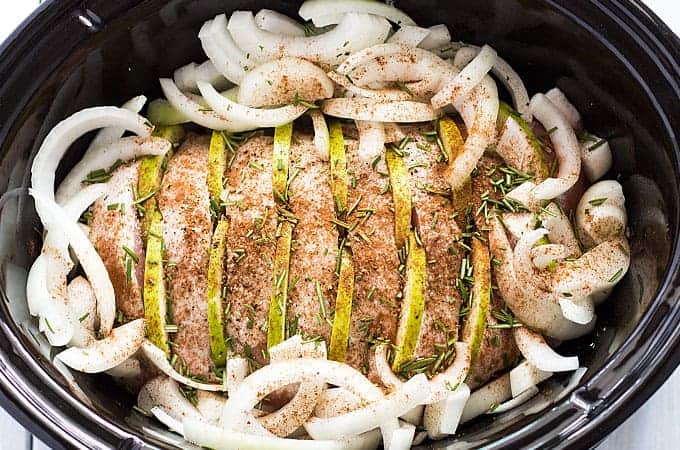 A raw seasoned pork loin stuffed with sliced apples and surrounded by sliced onion in a slow cooker.
