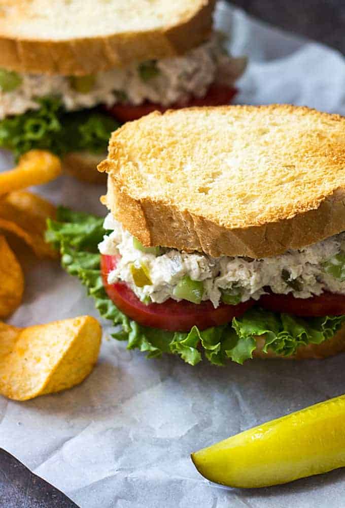 A closeup of a fish salad sandwich beside a pickle spear and chips.