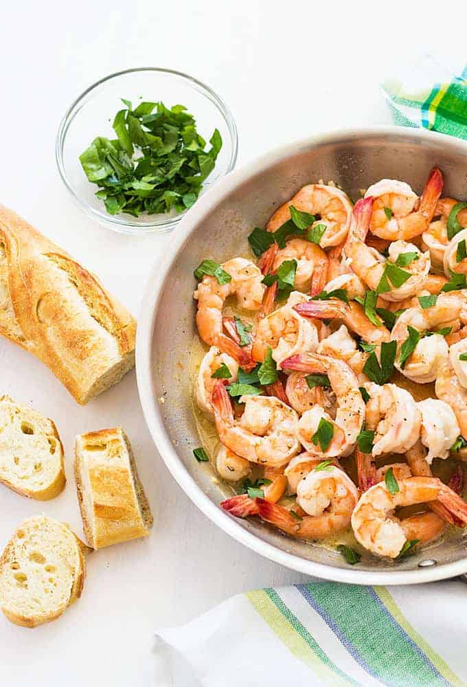 Overhead view of shrimp in a skillet beside a loaf of French bread and a bowl of parsley.