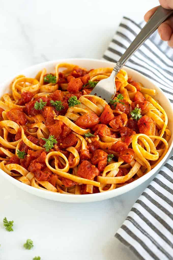 Pasta with tomato sauce topped with parsley in a white bowl with a fork beside a black and white striped napkin.