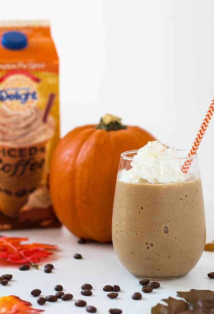 A coffee smoothie topped with whipped topping.  A pumpkin and carton of iced coffee is in background.