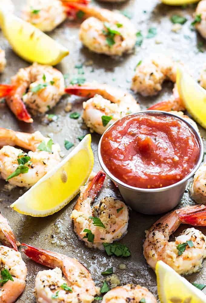 Baked garlic shrimp on a baking sheet with lemon wedges and a sauce cup of cocktail sauce.