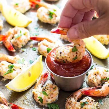 Baked shrimp with garlic and Parmesan on a baking sheet with a shrimp being dipped into a sauce cup with cocktail sauce